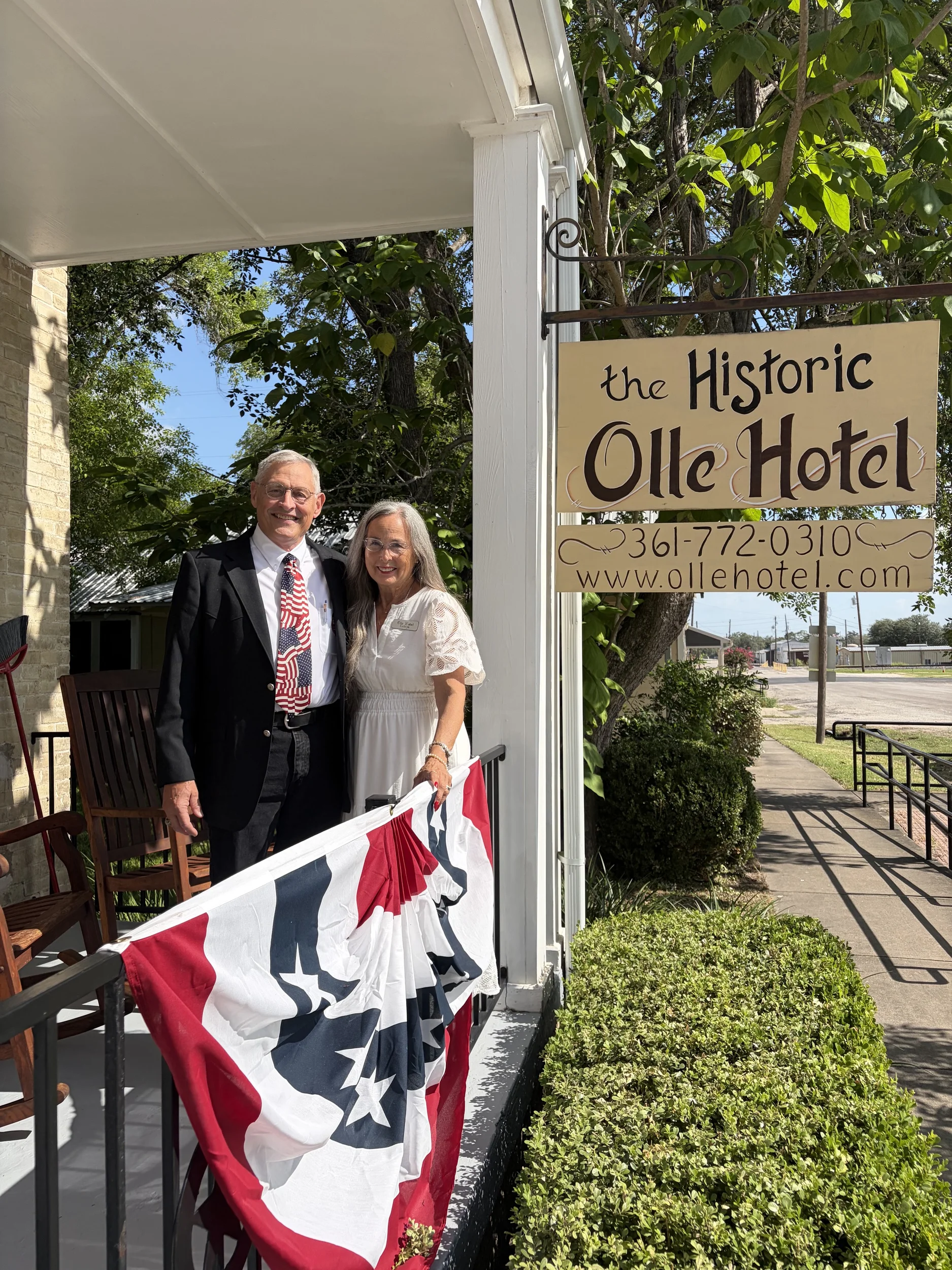 Two people posing on a porch beside a sign reading “Historic Olie Hotel,” holding a red-white-blue banner.